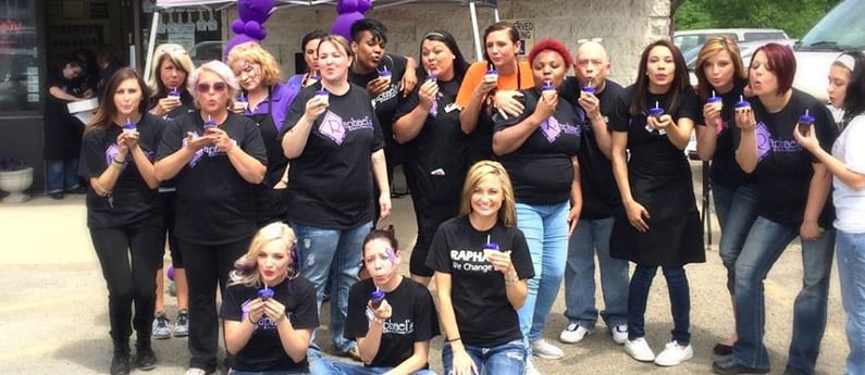 A group of Raphael’s School of Beauty students holding purple cupcakes with candles in the center and staff celebrating the school's anniversary